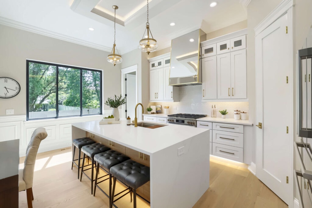 A bright, modern kitchen featuring white cabinetry, a large white quartz island with gold fixtures, black tufted barstools, and light hardwood floors. Photo by Virtuance.