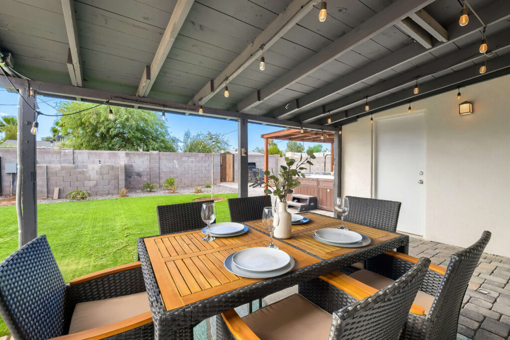 Staging exterior covered back patio highlighting an outdoor dining set with nice plate setting and a vase in the center of the table with greenery. Lawn in the background is nicel landscaped and in the back right you can see a hot tub and gazebo feature.