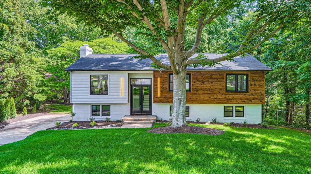 Exterior front view of a modern two-story split-level home featuring white siding, dark wood paneling, black trim windows, vertical light fixtures, and a lush green lawn framed by trees.