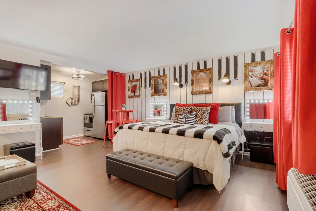A Virtuance real estate photograph of a uniquely styled studio apartment featuring a piano-key accent wall behind a king-sized bed. The space is decorated with bold red curtains, leopard print pillows, and ornate gold-framed wall art, with a small kitchen area visible in the background.