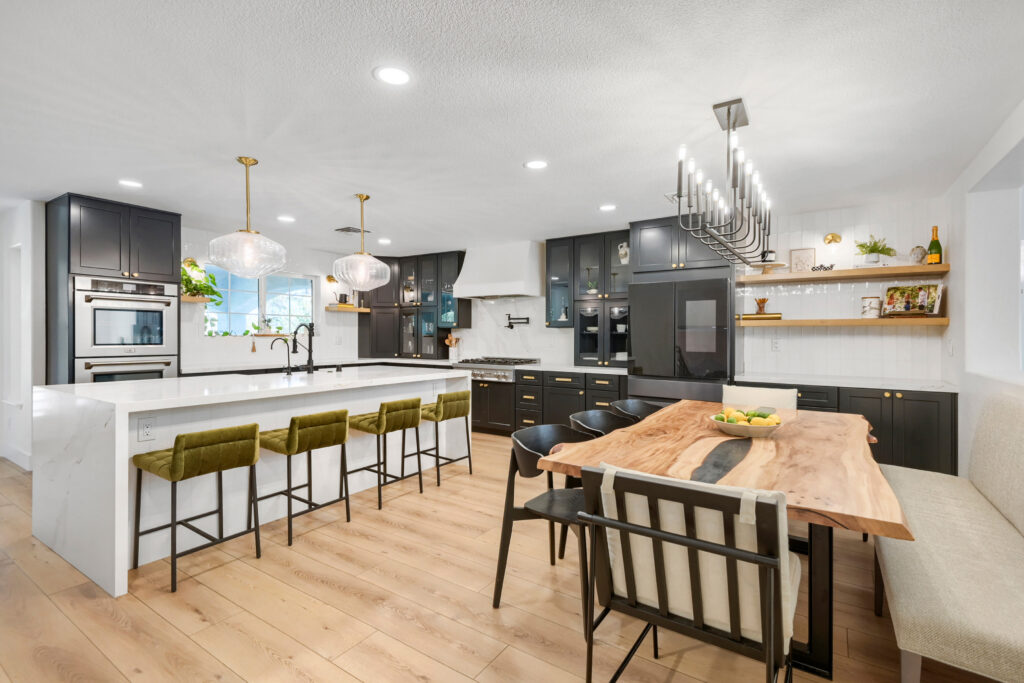 Modern open-concept kitchen and dining room featuring dark charcoal cabinetry, white quartz waterfall island with olive green barstools, and a live-edge wooden dining table beneath a linear chandelier.